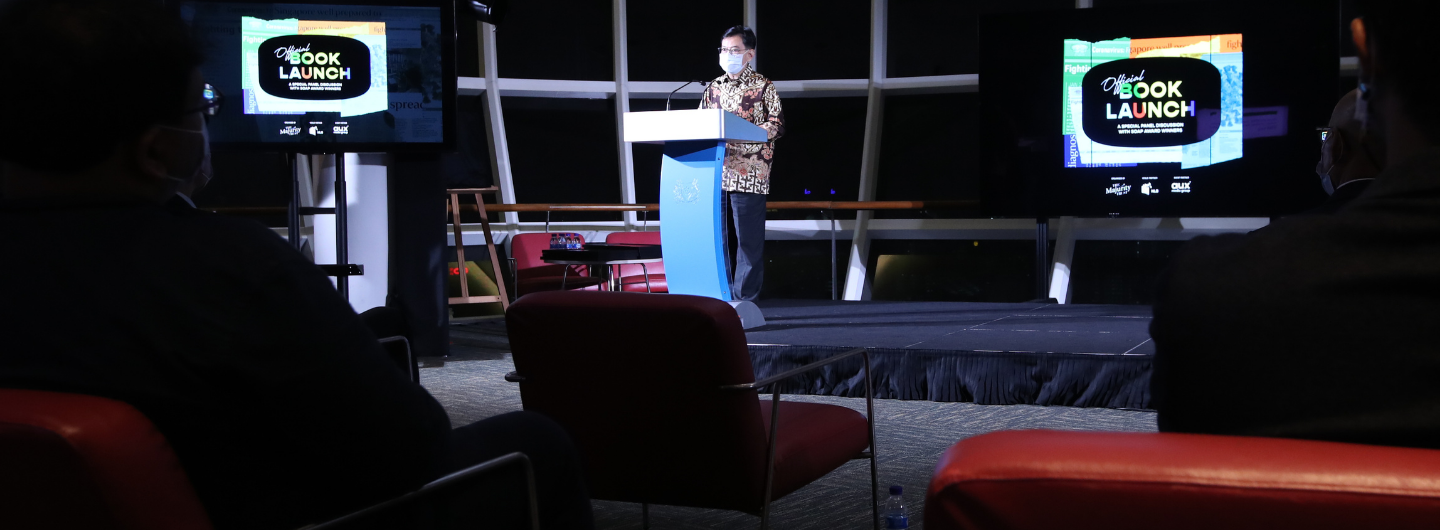 Man in mask at podium for "Official Book Launch." Monitors display event. Audience seated.